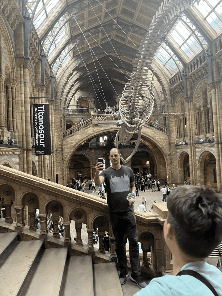 a man taking a selfie of a dinosaur skeleton on the stairs of the natural science museum in london